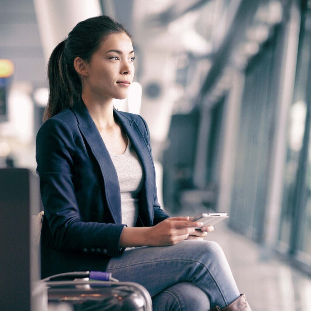 Mobile phone travel businesswoman waiting in airport lounge with hand luggage. Young Asian woman on business trip using cell smartphone. Tech device for wifi entertainment online in terminal.